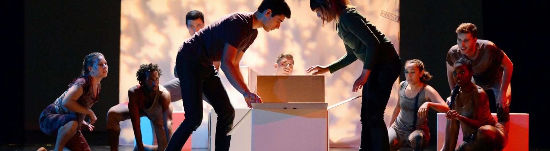 On stage, two actors look into an open box. An ensemble surrounds them.