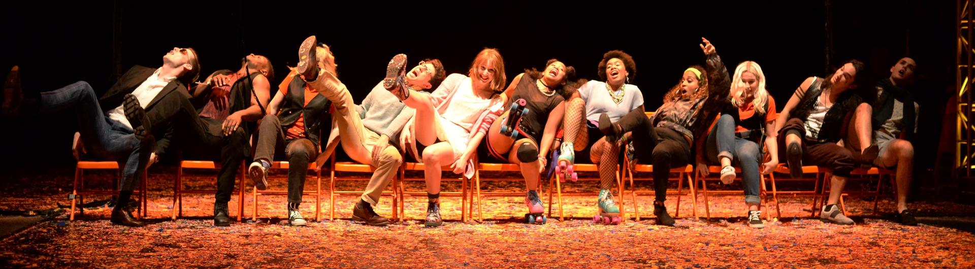 On stage, students in a line of chairs lean far to one side and have excited facial expressions