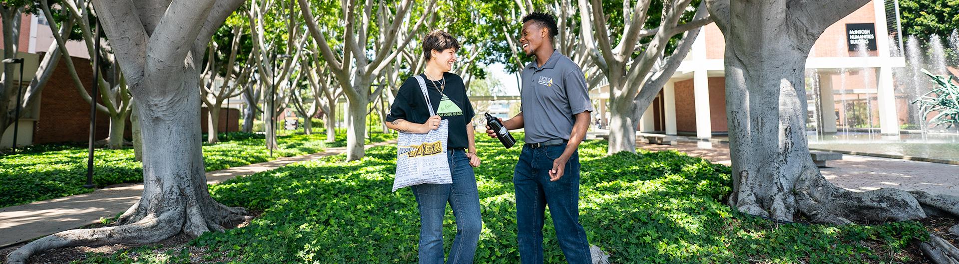 Two students wearing custom clothing and accessory items smiling at each other in front of green trees on the CSULB campus. 