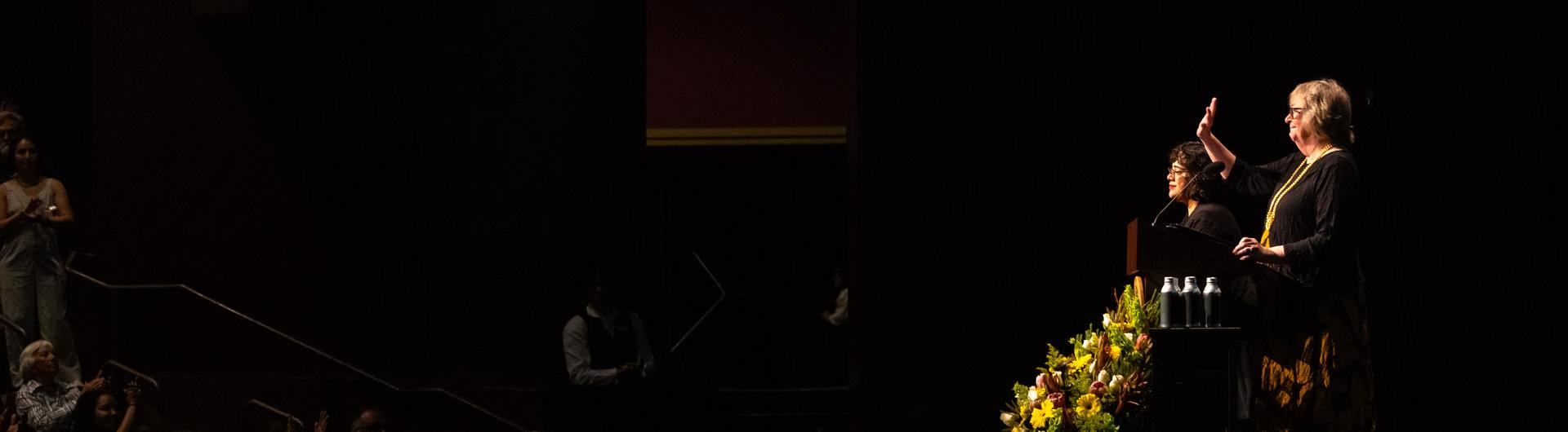 Campus President Jane Close Conoley, standing next to a sign language interpreter, waves while on stage during Convocation 2024