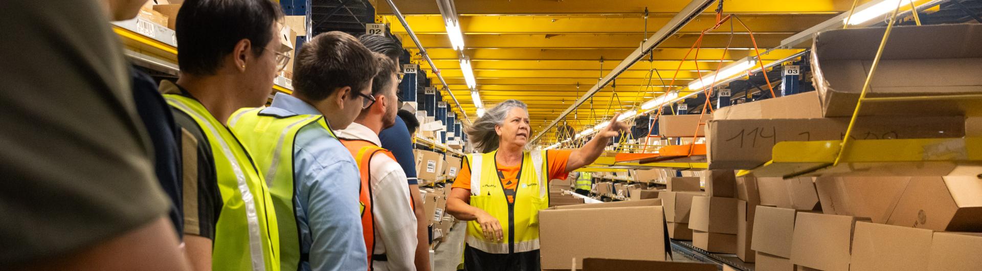 A tour guide explaining supply chain operations to students standing near a conveyer moving boxes