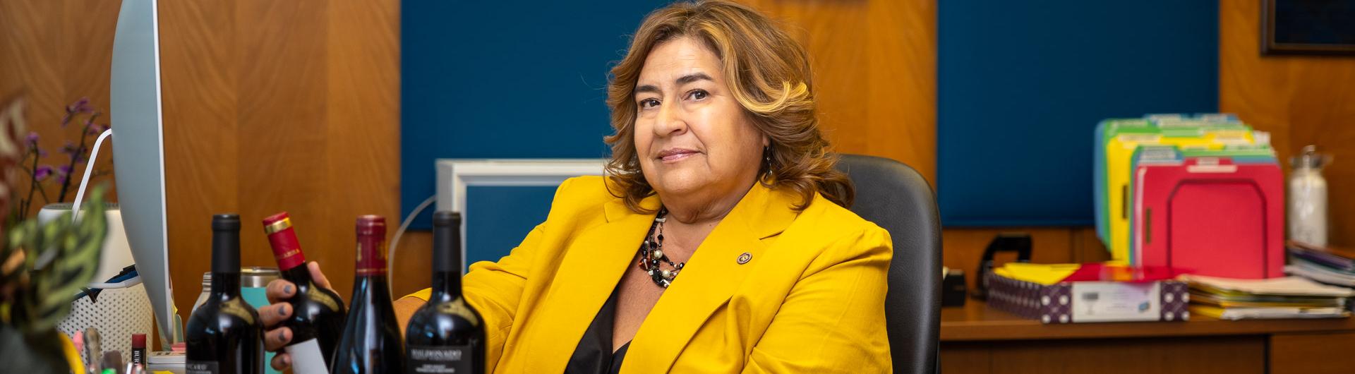 Anna Ortiz, in a yellow blazer, sits at an office desk with wine bottles, paperwork and colorful file folders in the background.