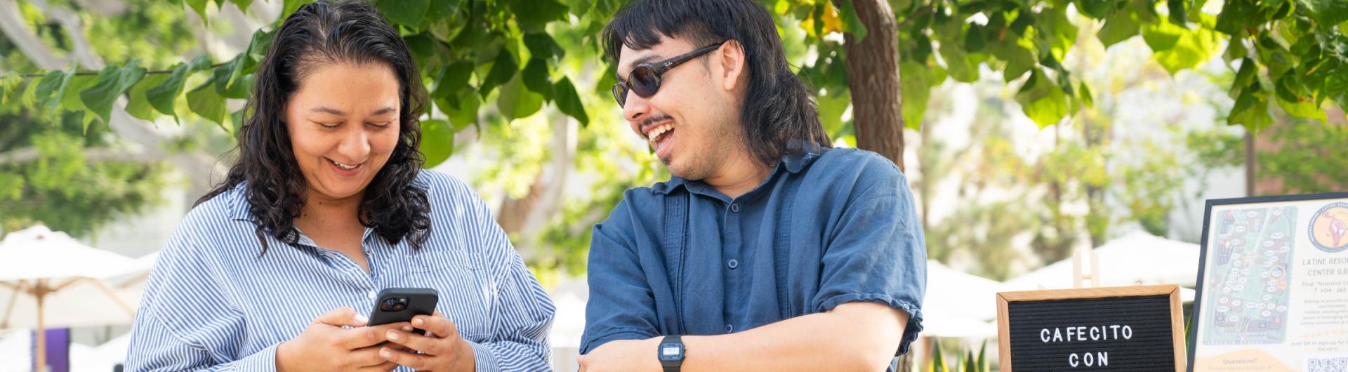 Two people standing together, one on their phone and one laughing.