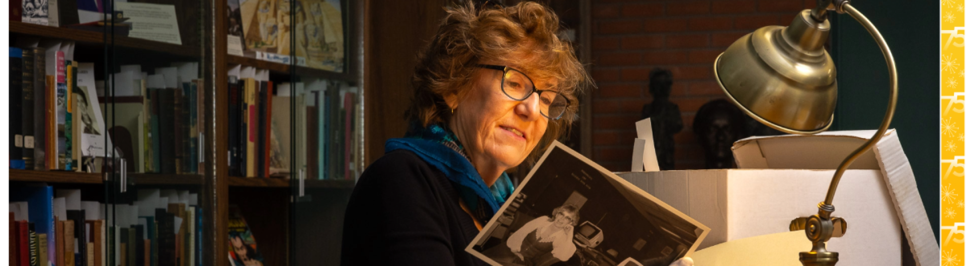 Under a desk lamp, author Barbara Kingsley-Wilson inspects an old black-and-white photo surrounded by archival boxes and documents.