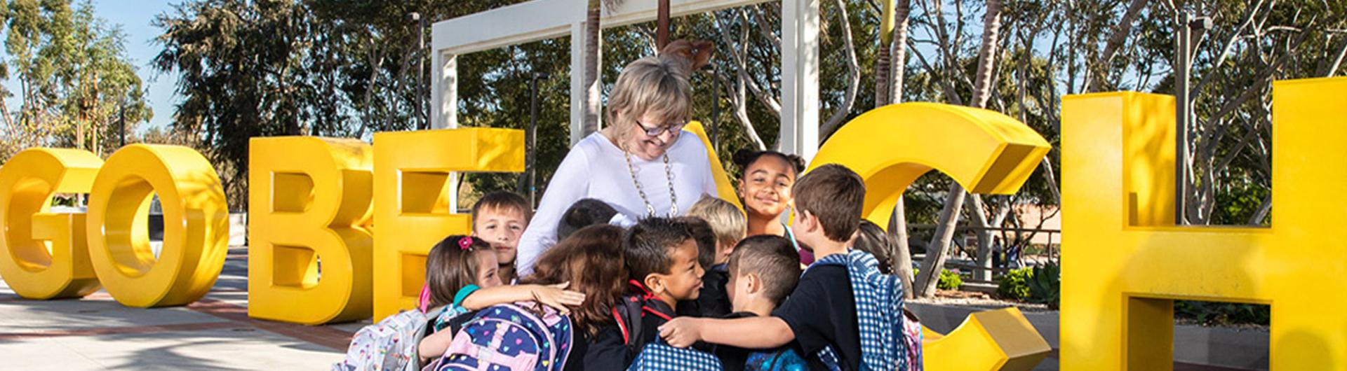 Long Beach State University President Jane Close Conoley stands among a group of children with backpacks who are hugging her in front of large yellow "GO BEACH" letters on campus.