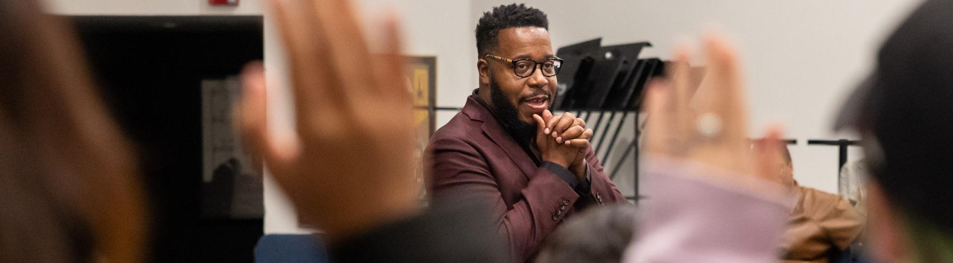 Ananias Montague, in a burgundy suit, speaks to a group with raised hands in the foreground.