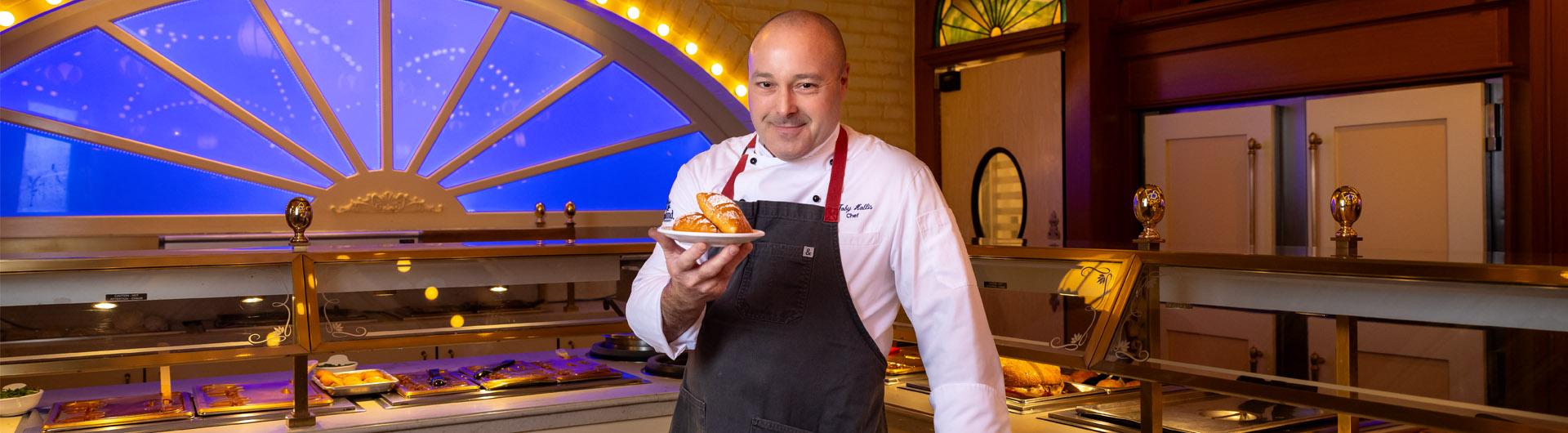 Disneyland chef and CSULB alumnus Toby Hollis holds a plate of beignets in front of a buffet with warm lighting.