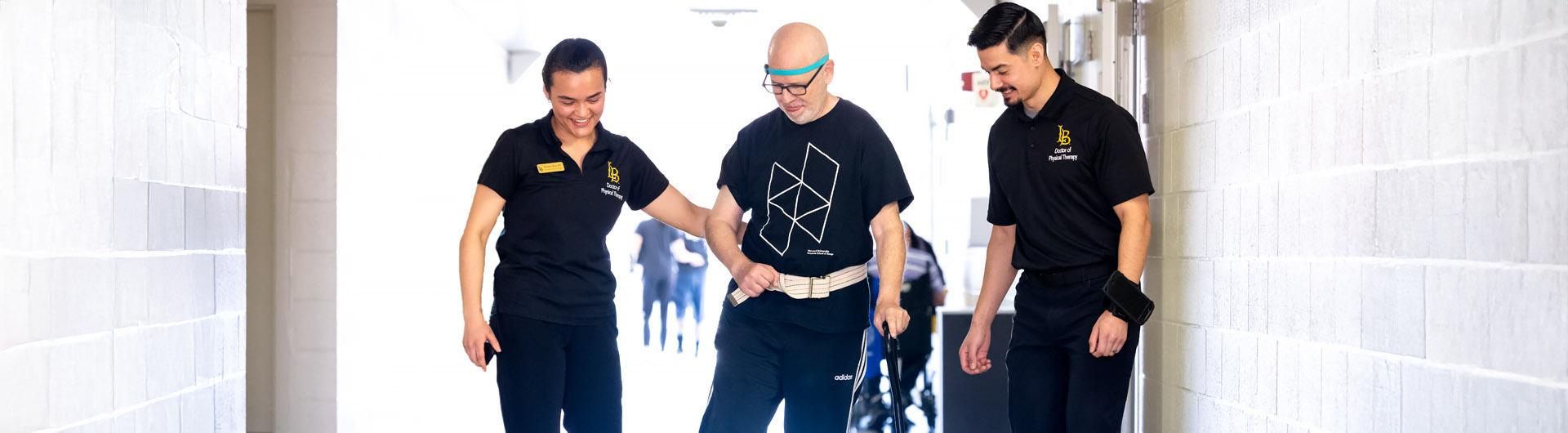 Two CSULB students walk beside a Neuro Lab client as he uses a cane to walk down a hallway.