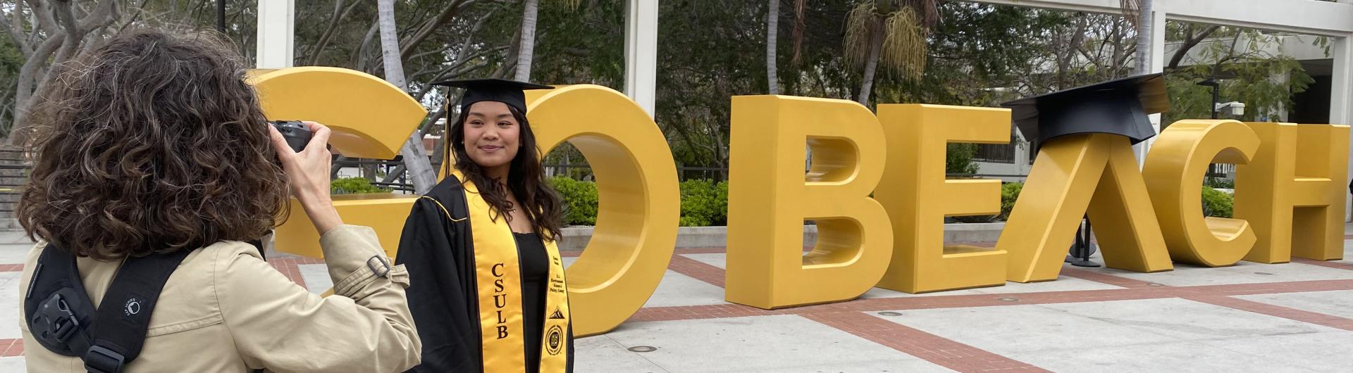 Student standing in front of the Go Beach sign wearing a cap and gown