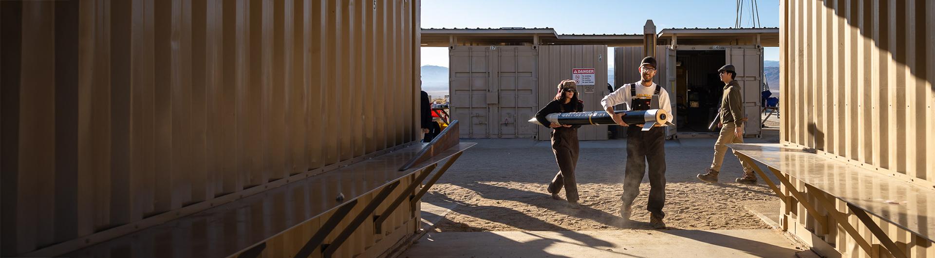 Three people carry a large rocket component at a desert facility
