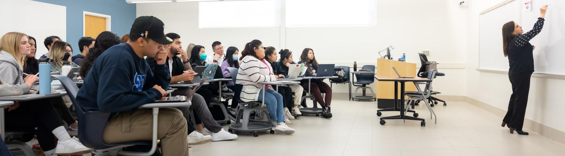 Professor writing on whiteboard with class of students watching.