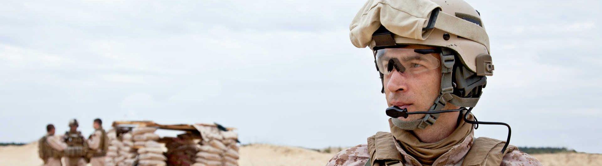 Three military men stand in front of a sandbag shelter while another man in military gear gazes over the desert