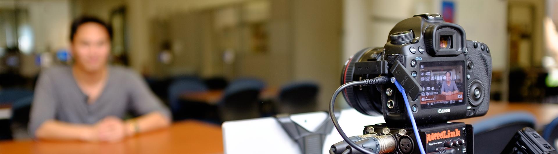 A student sits before a camera with a teleprompter.