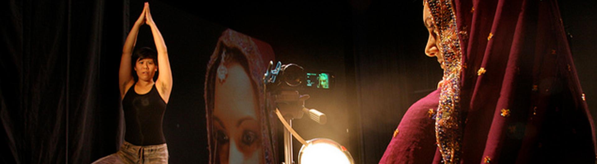 Shyamala Moorty performing on stage with another dancer while she is on camera and projected on a rear screen.