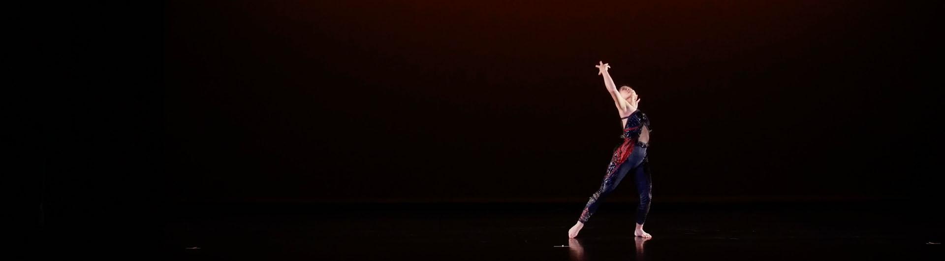 A dancer stands on a dark stage looking towards the sky with her hands raised up.