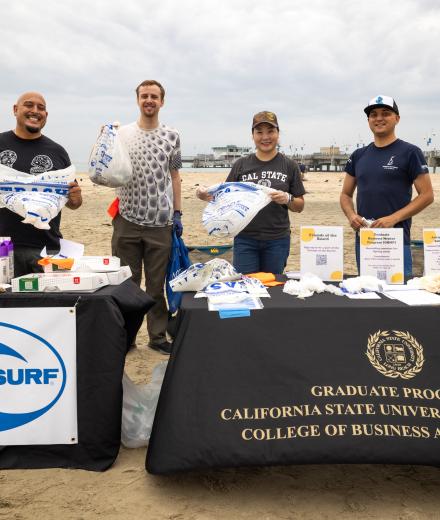 A GROUP OF PEOPLE STANDING BEHIND TABLES WITH ITEMS TO COLLECT TRASH AT THE BEACH