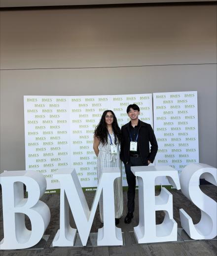Woman with dark long hair and light outfit standing next to man with dark short hair and dark outfit; both behind letters that say "BMES"