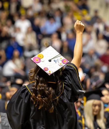 An image of a person in a cap and gown from behind with their hand rasied towards a crowd of people.