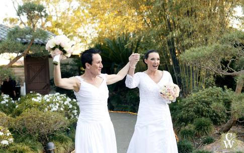 Two brides walk down the entrance path into the Japanese Garden, smiling.