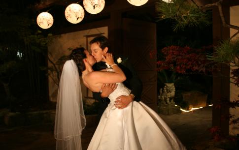 A bride and groom kiss outside the gate of the Japanese garden at night. Lanterns are lit in the background.