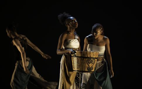 Three dancers on stage with a bucket of water and costumes including Adinkra symbols and cowrie shells.