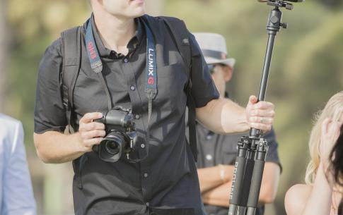 Gregory Crosby dressed all in black holding two cameras, one around his neck and one on a tripod. The background has a few out of focus people against distant trees.