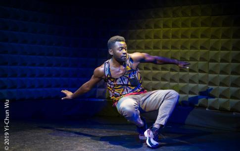 T performs on a dark stage illuminated by a spot light in grey sands and a colorful tank top.