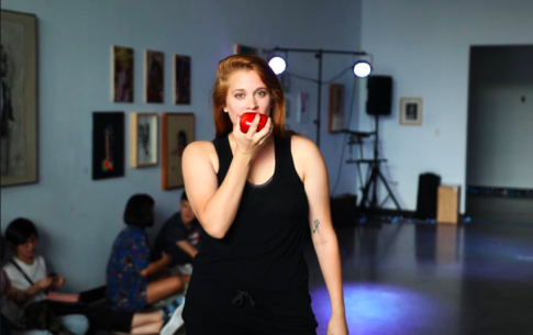 AJ Sharp dressed in a black tank top begins to bite into an apple while in a dance studio.