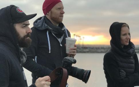 Gregory R.R. Crosby is wearing a baseball cap and sweater while manning a camera and standing next to two grad students who are also bundled up in jackets with a sunrise behind them.