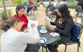 three students sitting down and painting on canvas 