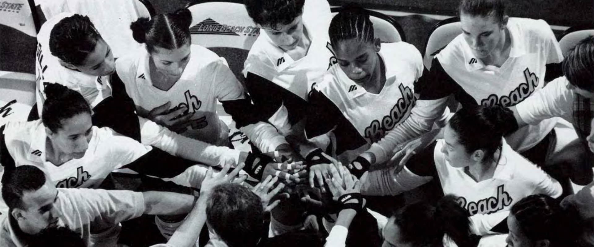 A women's volleyball team huddles in a circle with their hands together in a show of unity before a game.