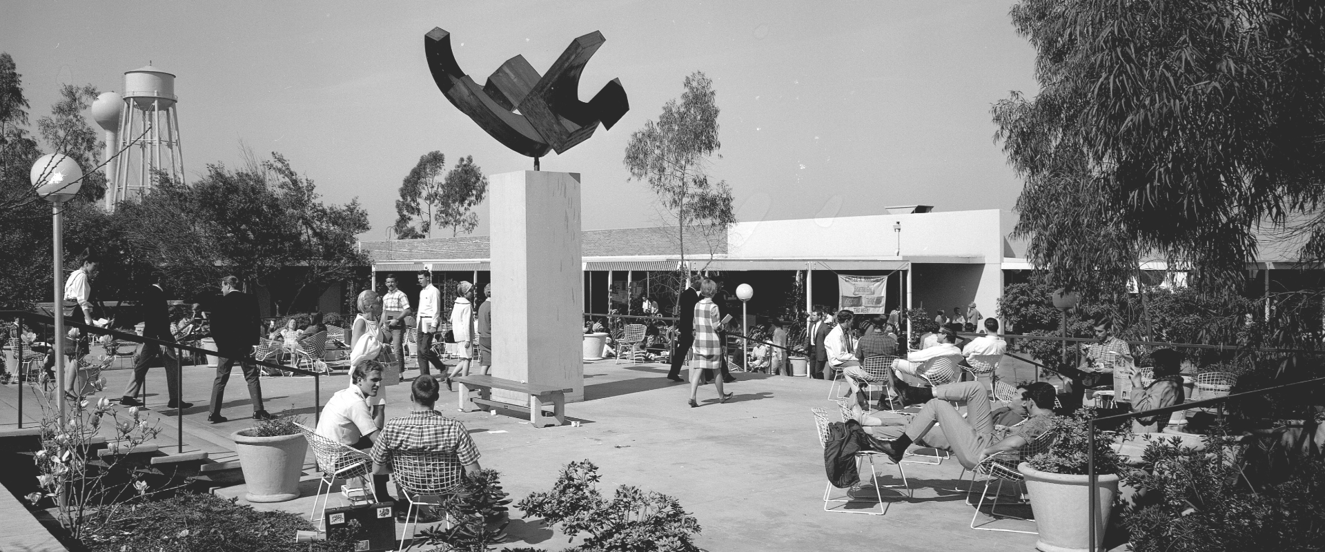 People gather and relax on a busy patio near a sculpture, with trees and a water tower in the background.