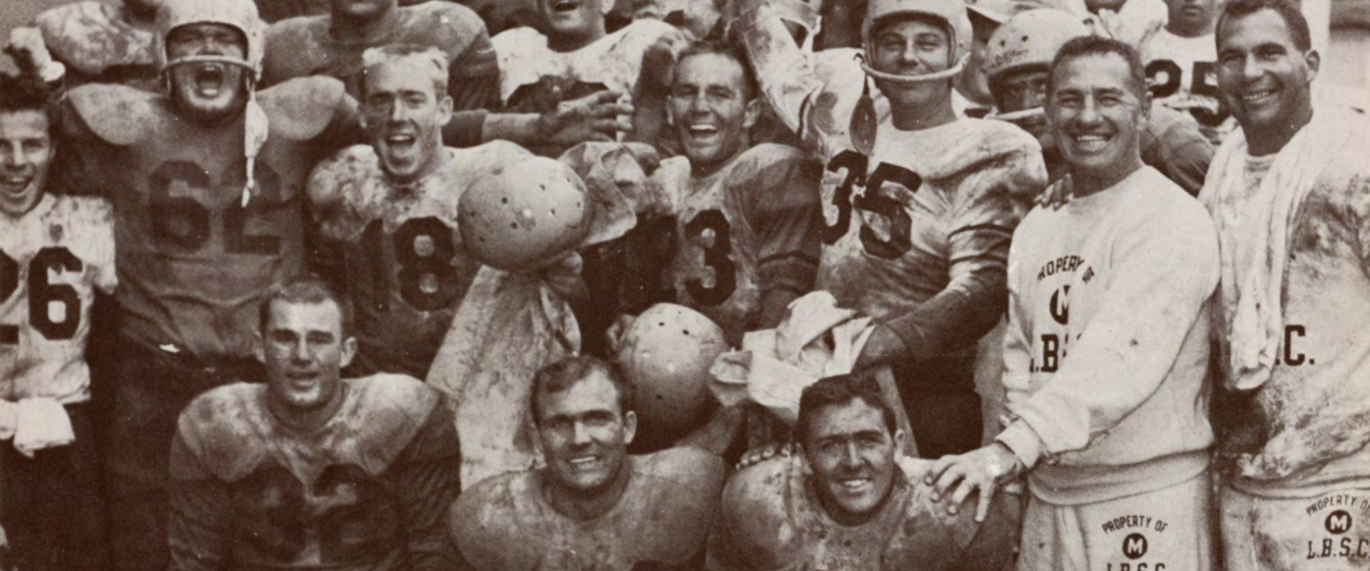 A muddy football team poses together, smiling and holding helmets after a game.
