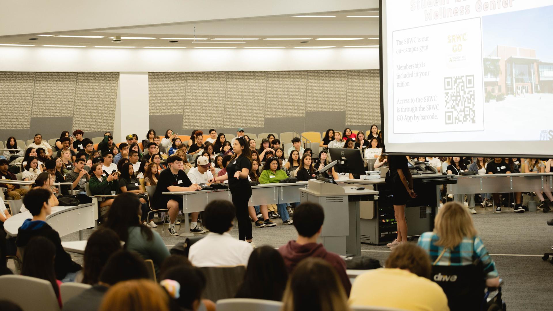 "A large group of students seated in an auditorium, listening to a presenter in front of a projection screen displaying information about the Student Recreation & Wellness Center.