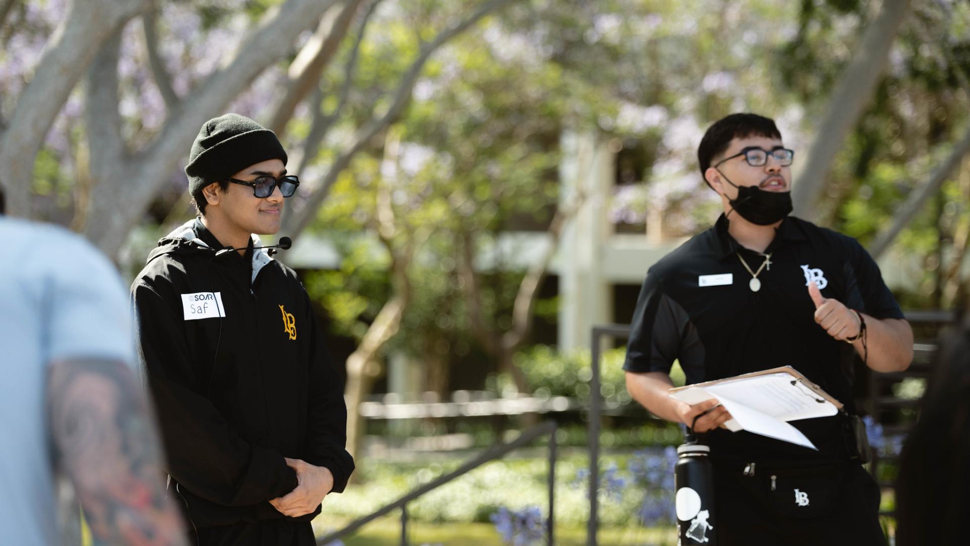 Two student leaders standing outdoors, one wearing a black beanie and sunglasses with a microphone headset, and the other holding a clipboard and giving a thumbs-up.