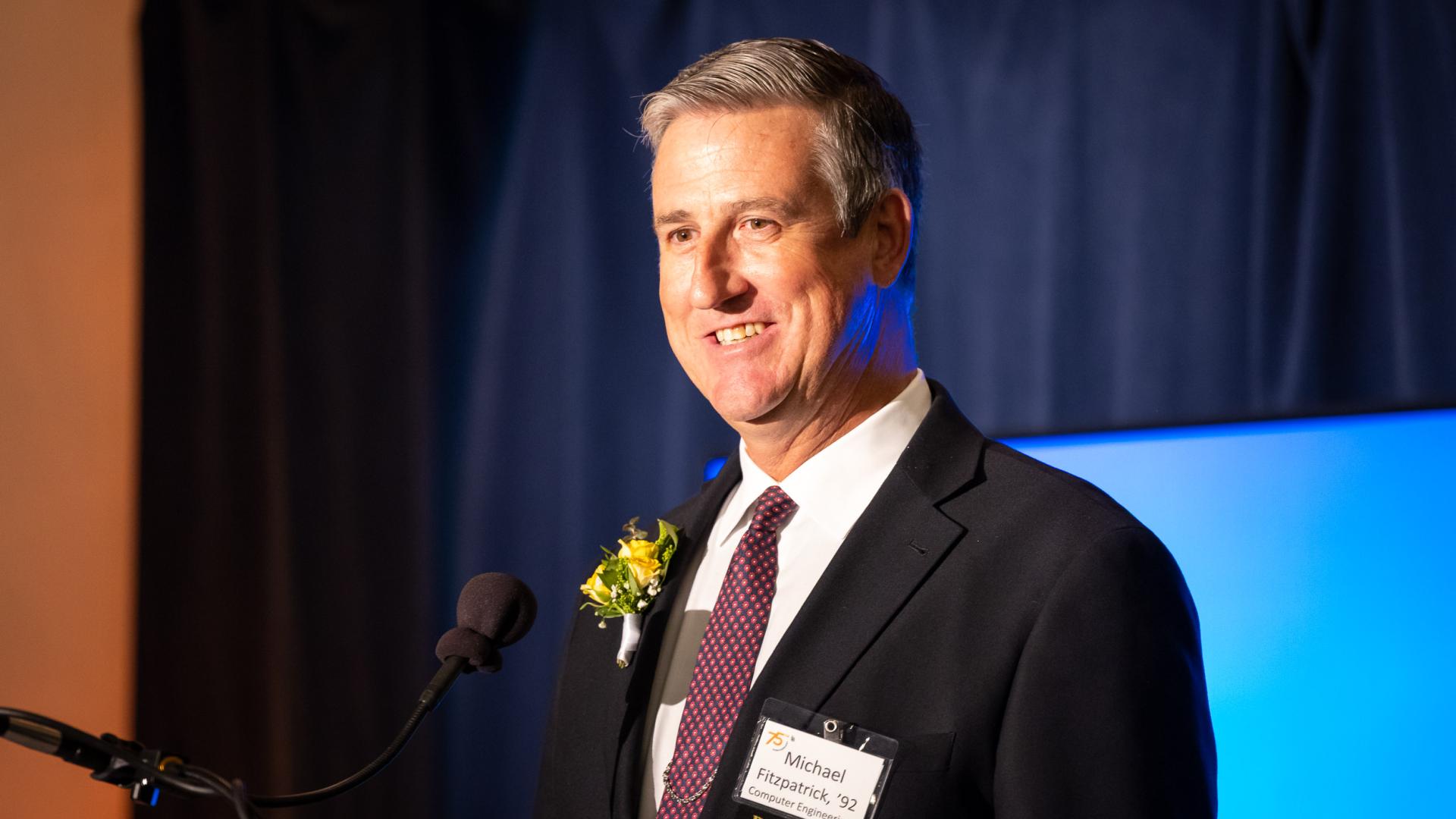 Distinguished Alumni honoree Michael Fitzpatrick smiles while standing behind a microphone