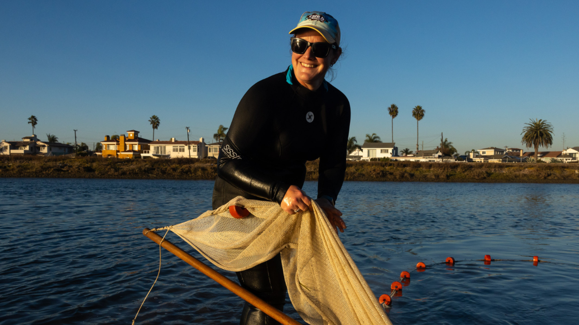 A woman in a wetsuit holds a net by a body of water