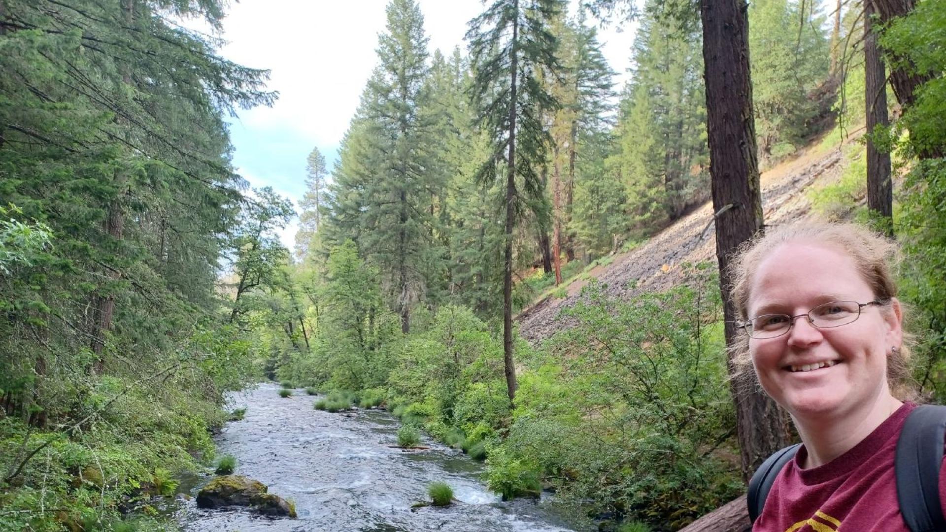 A woman stands with a river and forest in the background