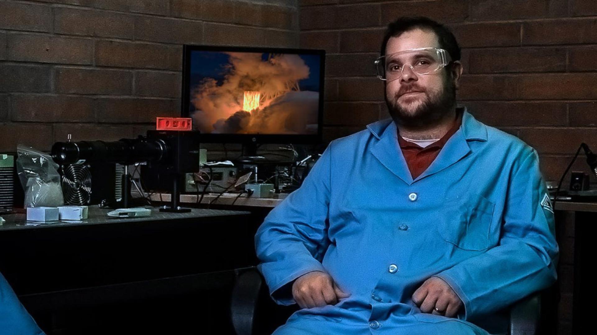 A man in a blue lab coat sits next to computer screen showing combustion