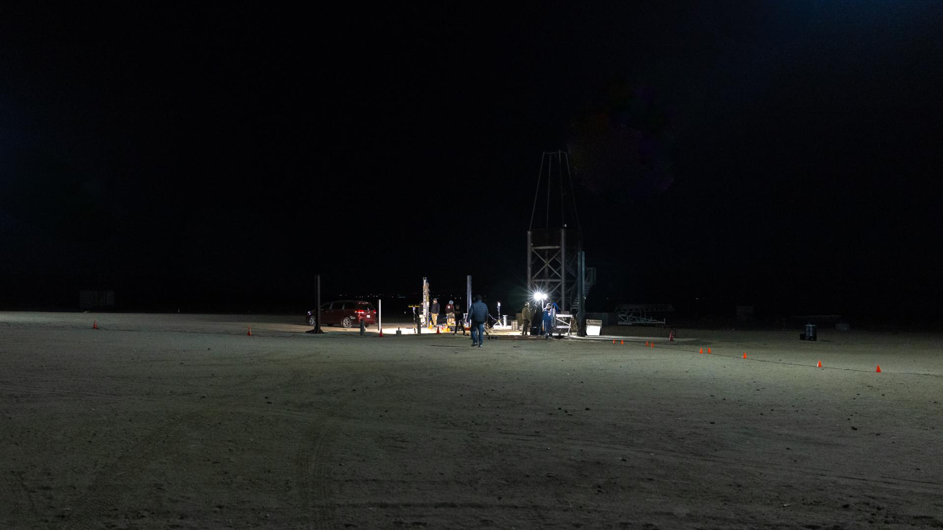 A group of CSULB students gather around a lit rocket test site in the dark