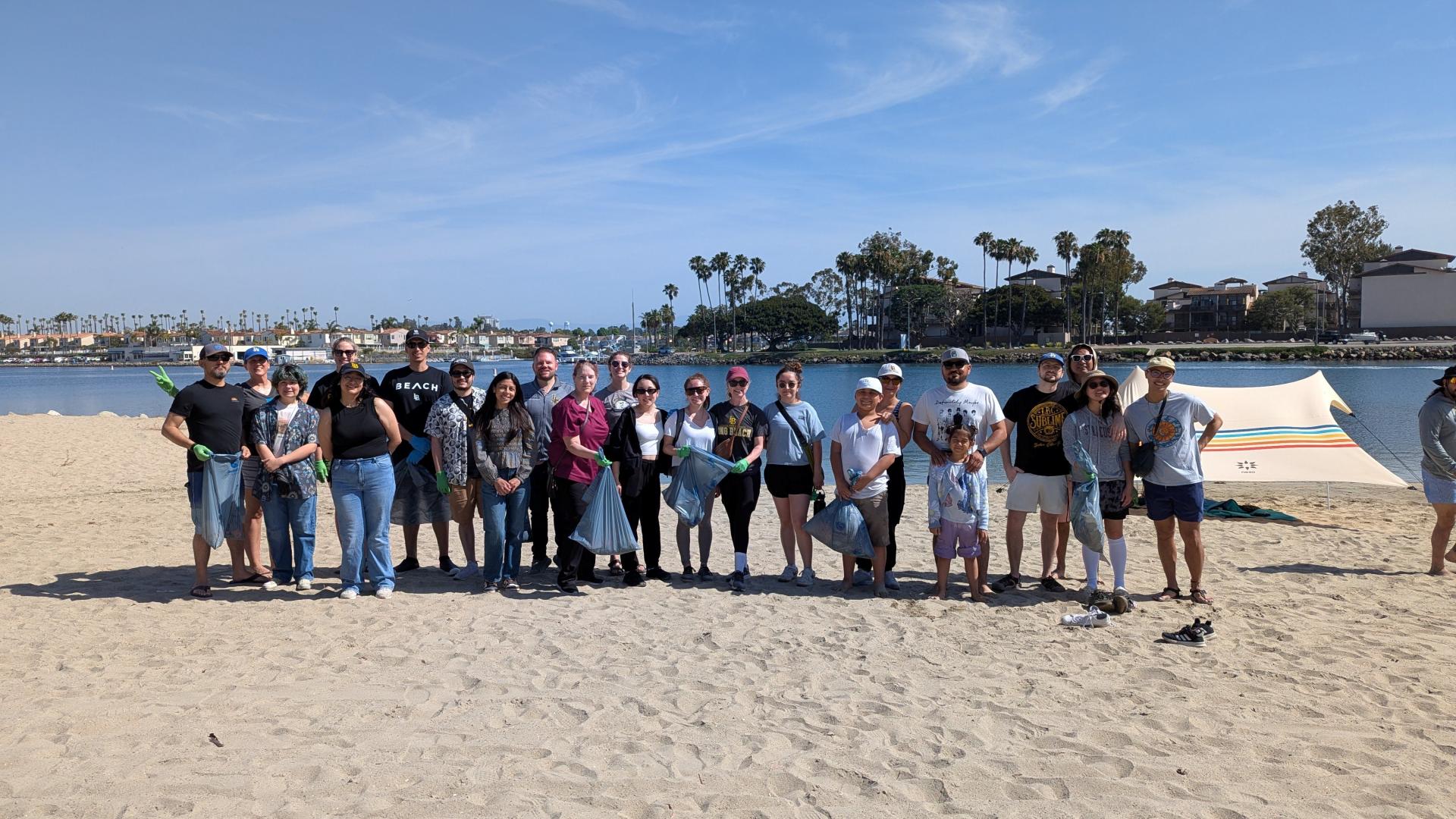 Beach cleanup volunteers pose with trash bags on a sunny shoreline with palm trees and water in the background