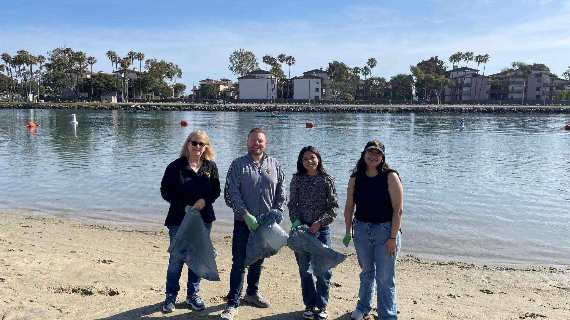 Volunteers wearing gloves hold trash bags during a beach cleanup near water and palm trees