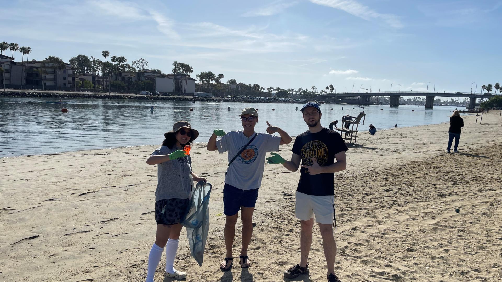 Volunteers wearing gloves collect trash during a beach cleanup near calm water and a bridge
