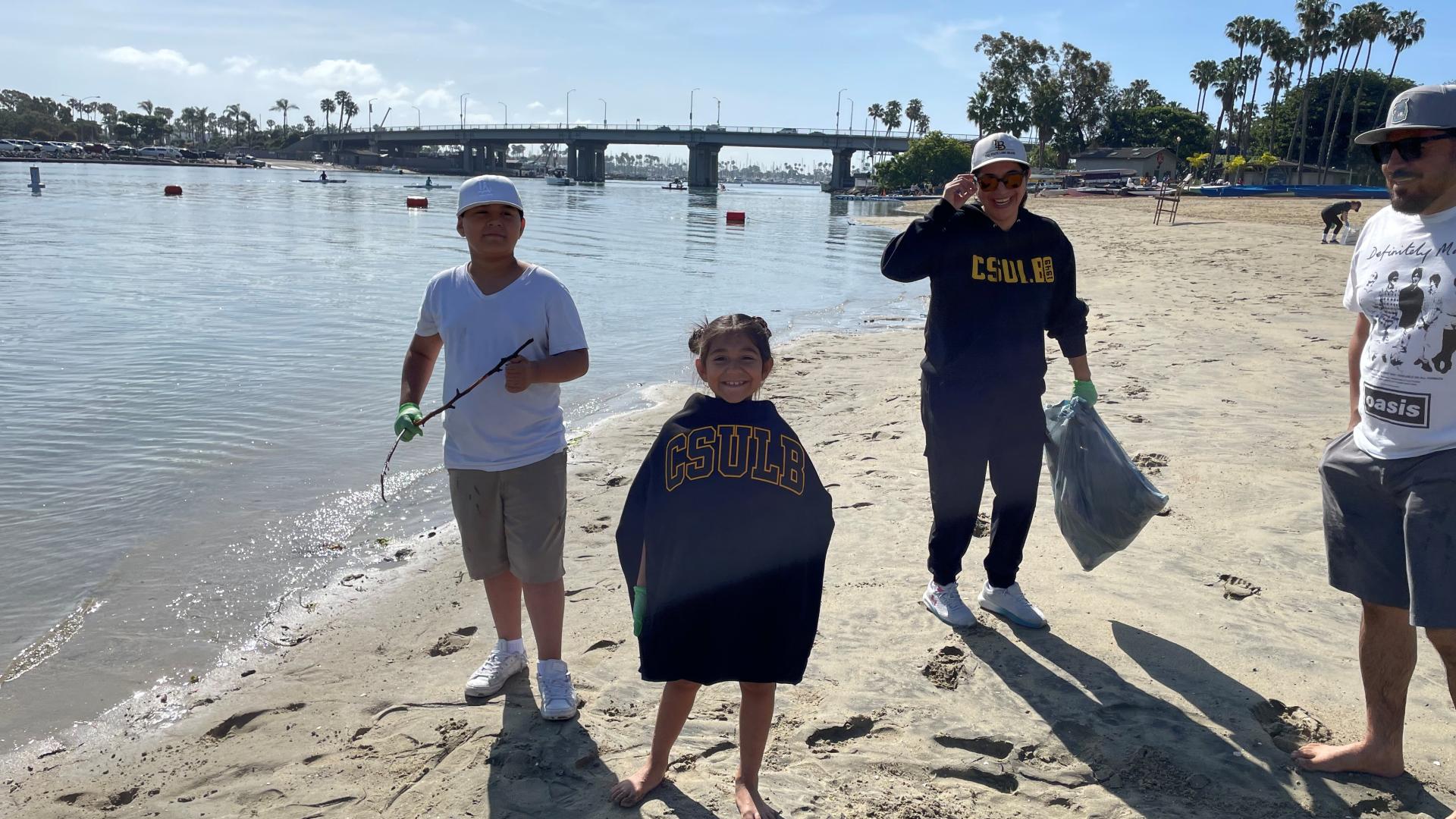 Two children and adult volunteers wearing gloves participate in a beach cleanup near the water and bridge.