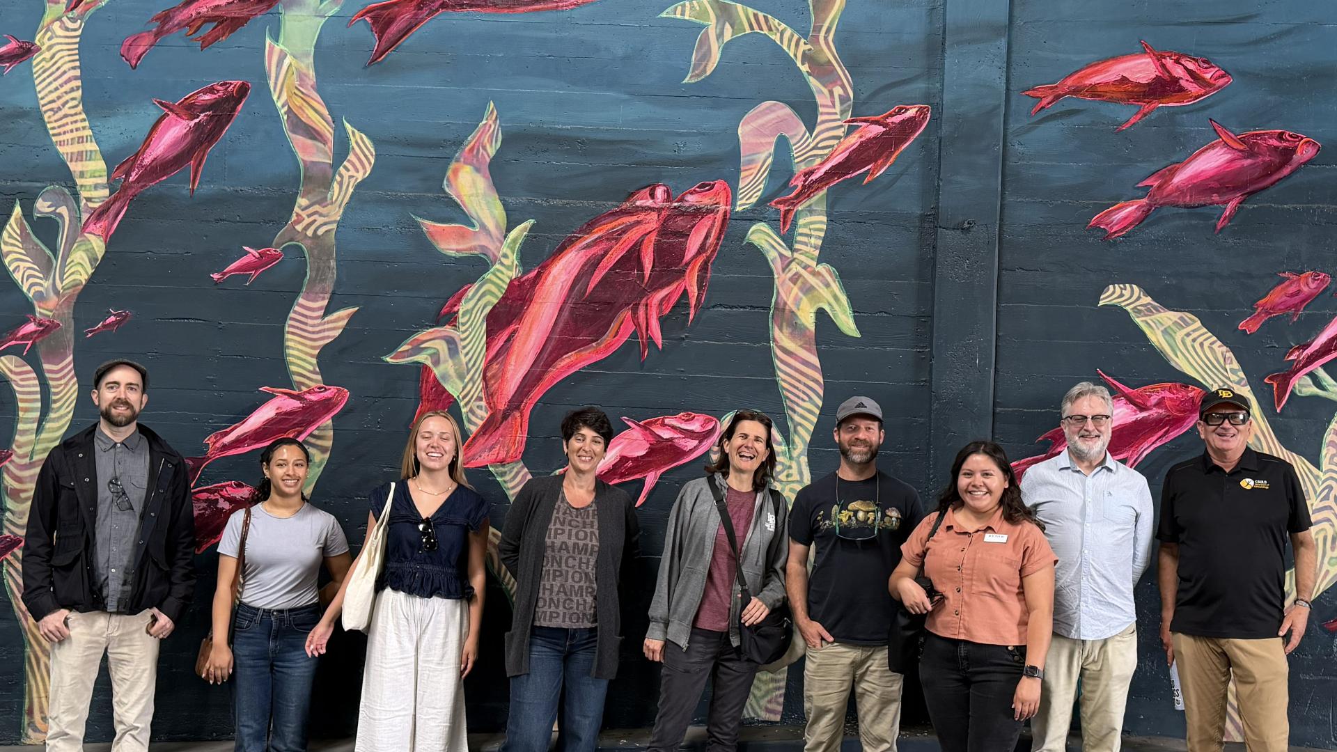 A group of people standing in front of a mural with red fish and green seaweed