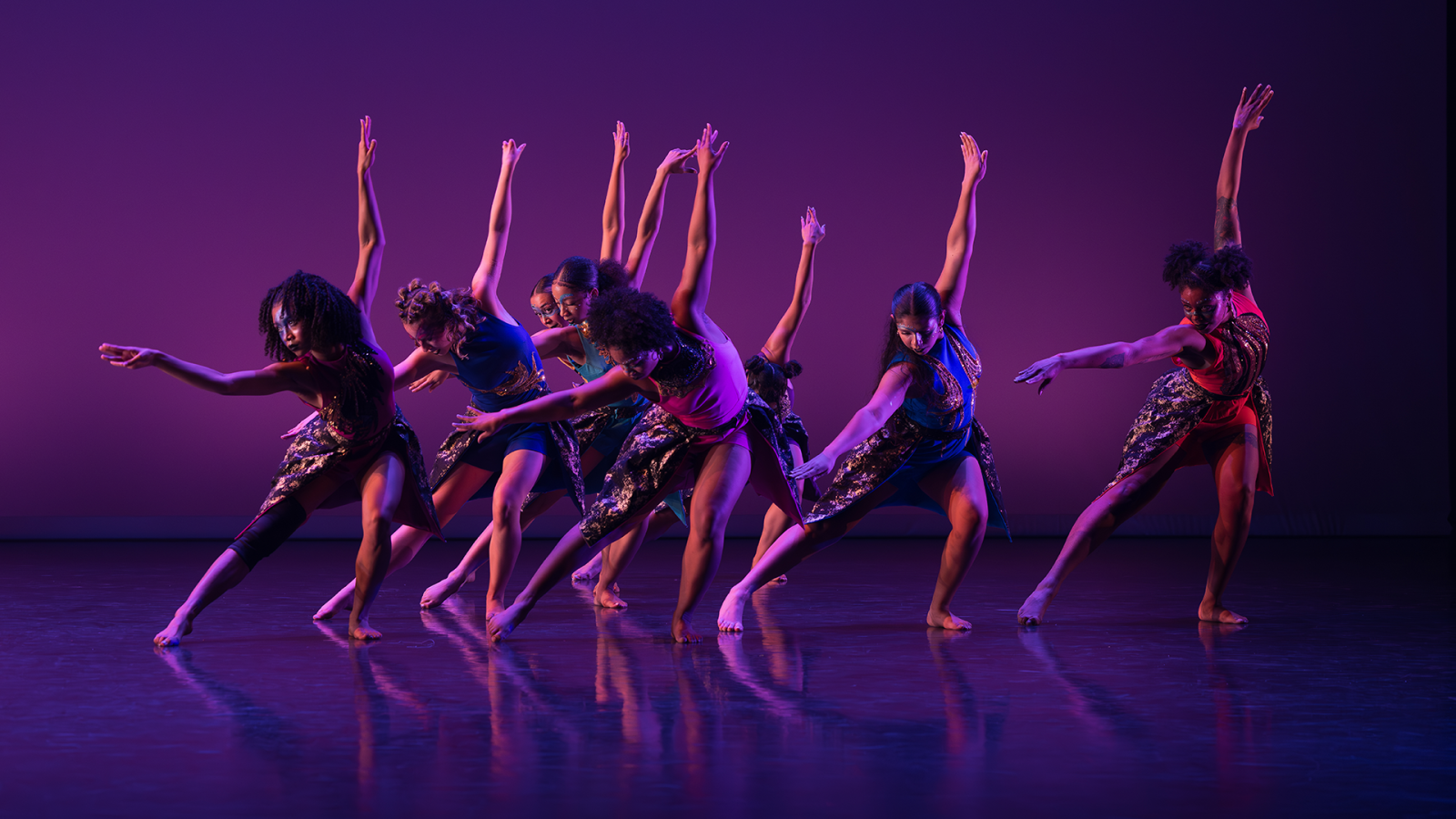 Eight dancers, awash in purple and pink light, reach out their hands on stage.