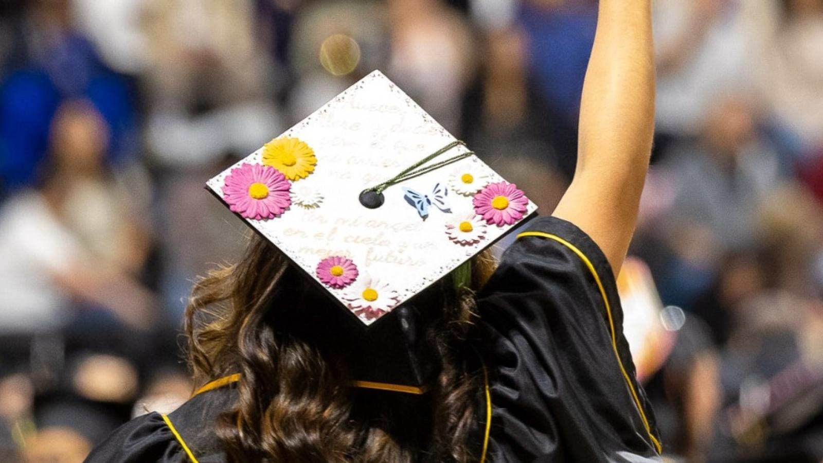 An image of a person in a cap and gown from behind with their hand rasied towards a crowd of people.