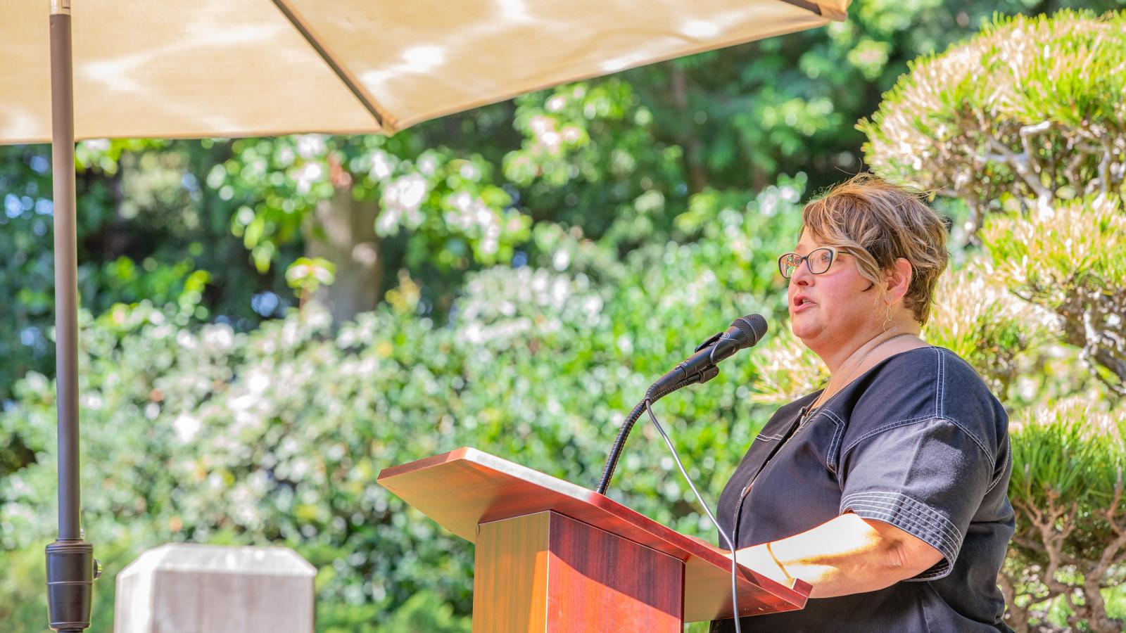 Professor Beth Manke speaking at podium under an umbrella