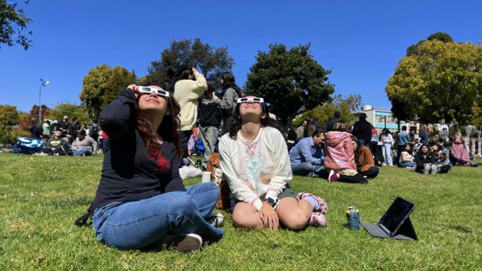 people sitting on grass watching eclipse with viewing glasses