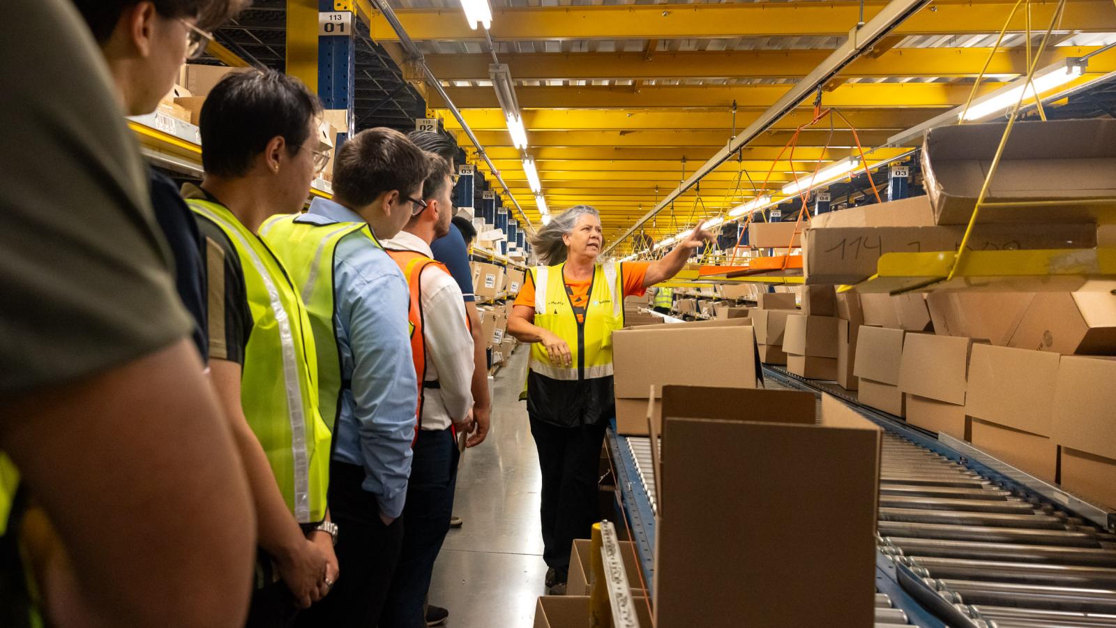 A tour guide explaining supply chain operations to students standing near a conveyer moving boxes